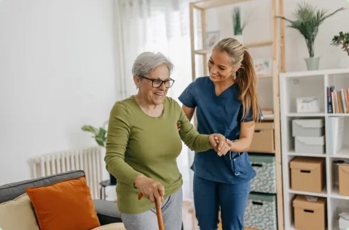 Caregiver helping an elderly woman with a cane walk across her room