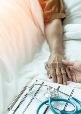 Caregiver gently holding the hand of a resident resting in bed, beside a clipboard and stethoscope